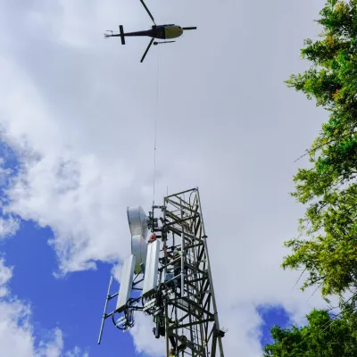 a helicopter flying in the sky with a cell tower on a long line external load