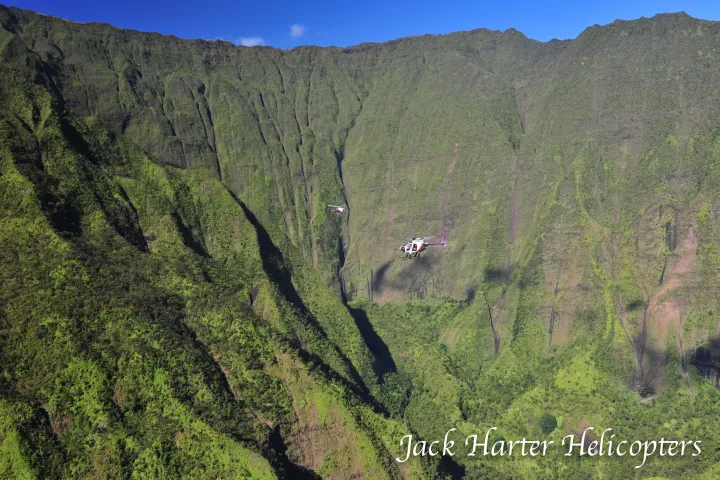 Helicopter flying over lush green mountains with deep valleys and a clear blue sky.
