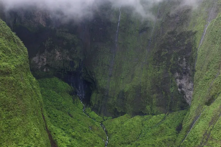 Lush green canyon with a narrow waterfall under cloudy skies.
