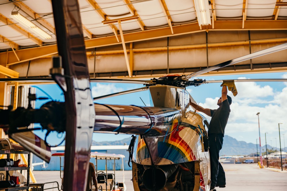 Person inspecting helicopter rotor inside a hangar with mountains in the background.