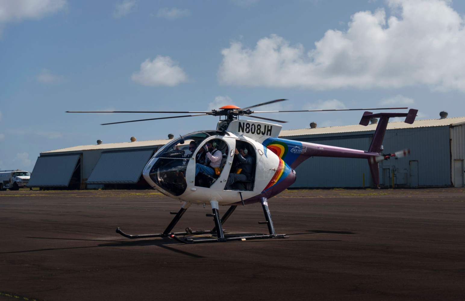 White helicopter on tarmac with hangars in the background under a partly cloudy sky.