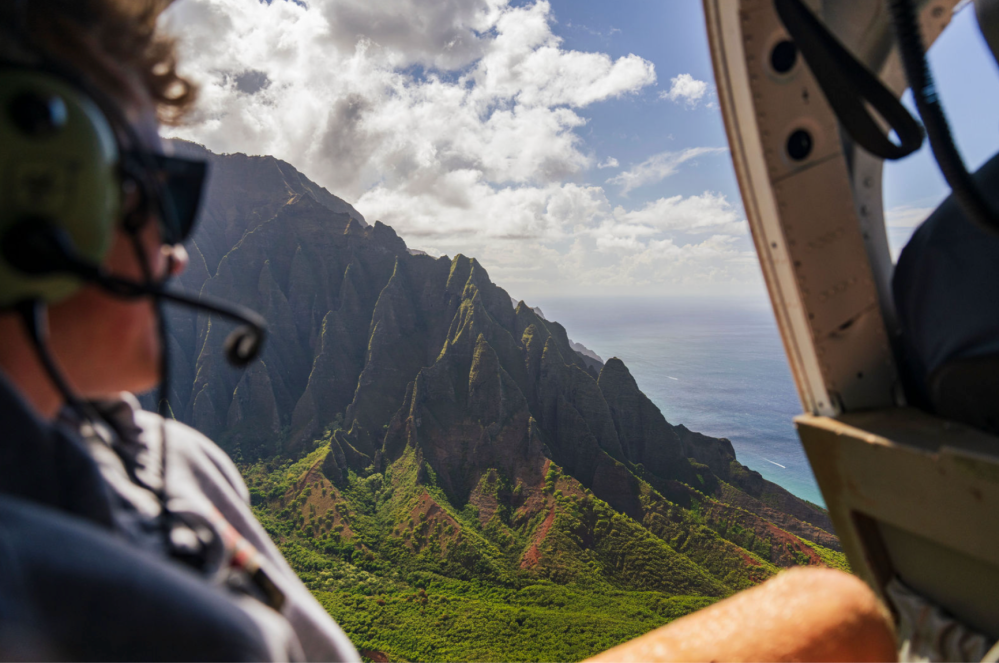Person in helicopter looking at ocean and mountains through open door.