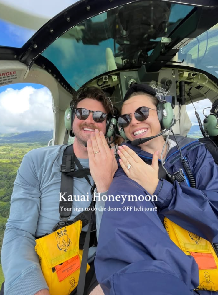 Two people in a helicopter smiling, showing rings, with life vests labeled 'Kauai Honeymoon' in view.