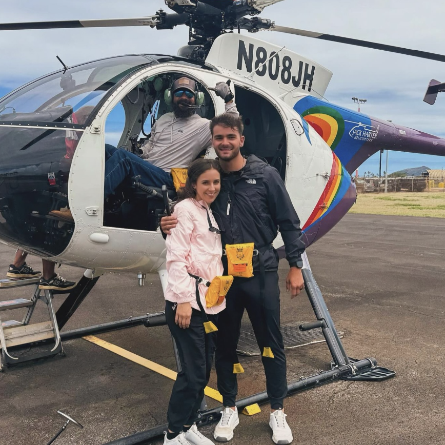 Smiling couple poses by a colorful helicopter with pilot inside waving.