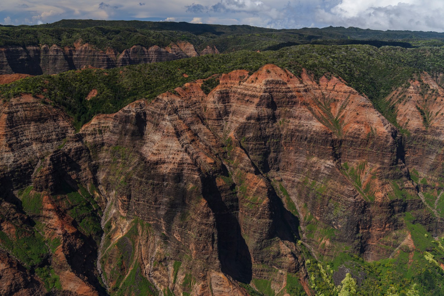 Aerial view of a rugged, green and red canyon landscape under a cloudy sky.