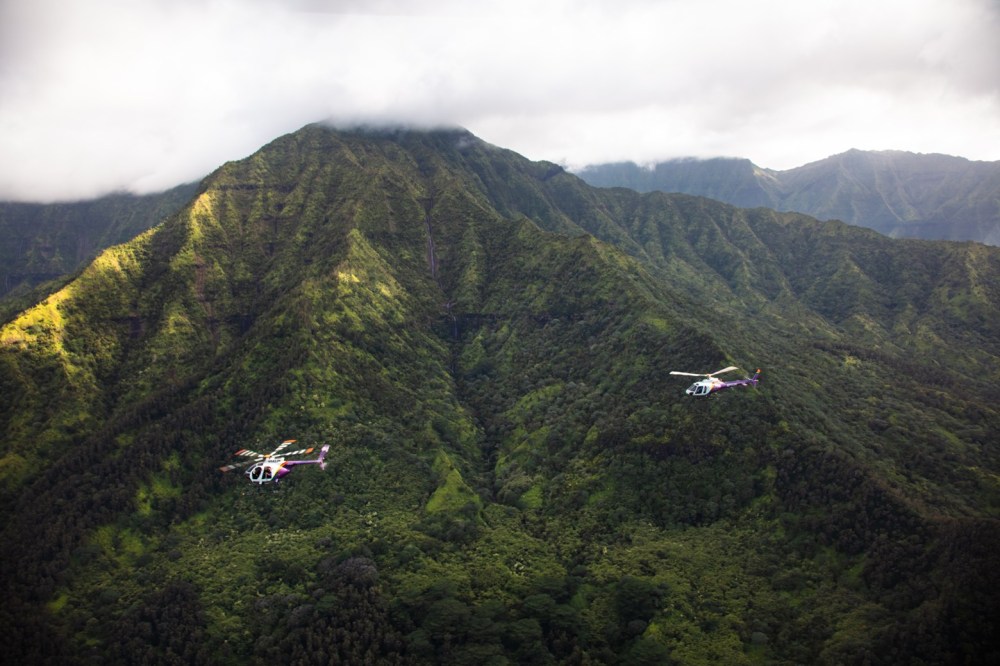 Two helicopters flying over lush green mountains with cloud-covered peaks.