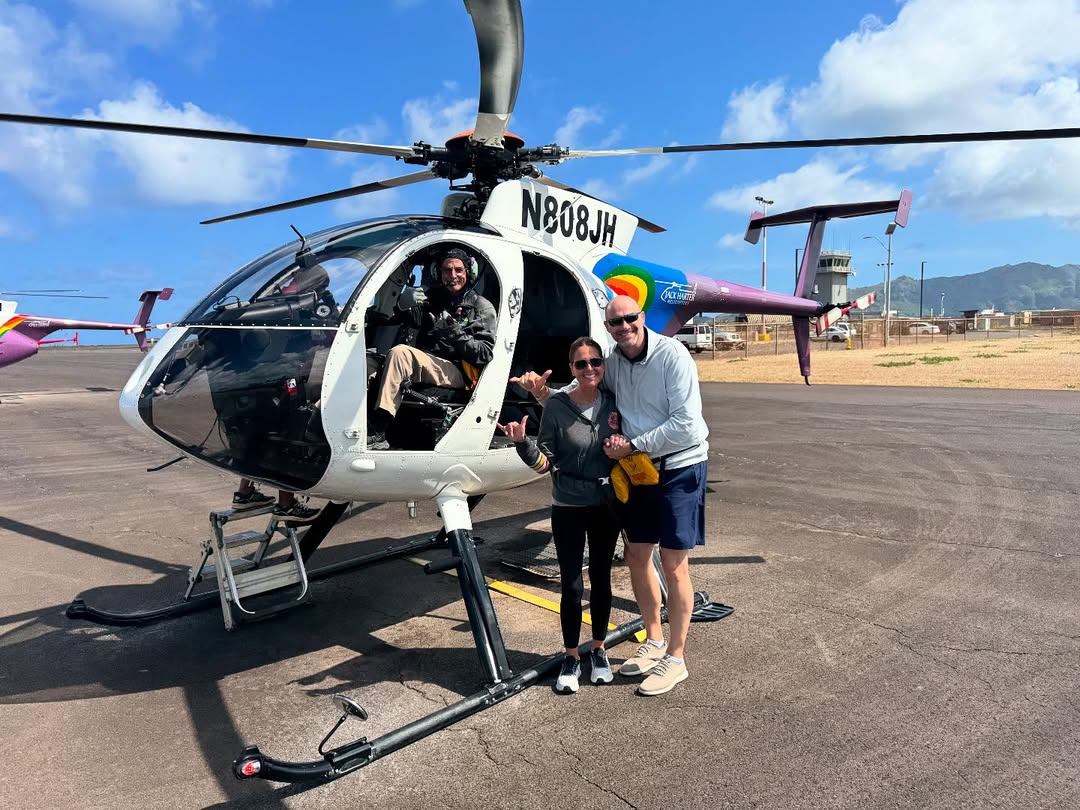Two people stand next to a helicopter, pilot sitting inside, on a sunny day.