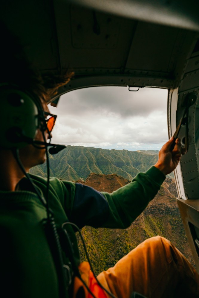 Person wearing headset inside helicopter, looking at lush green mountains through open door.