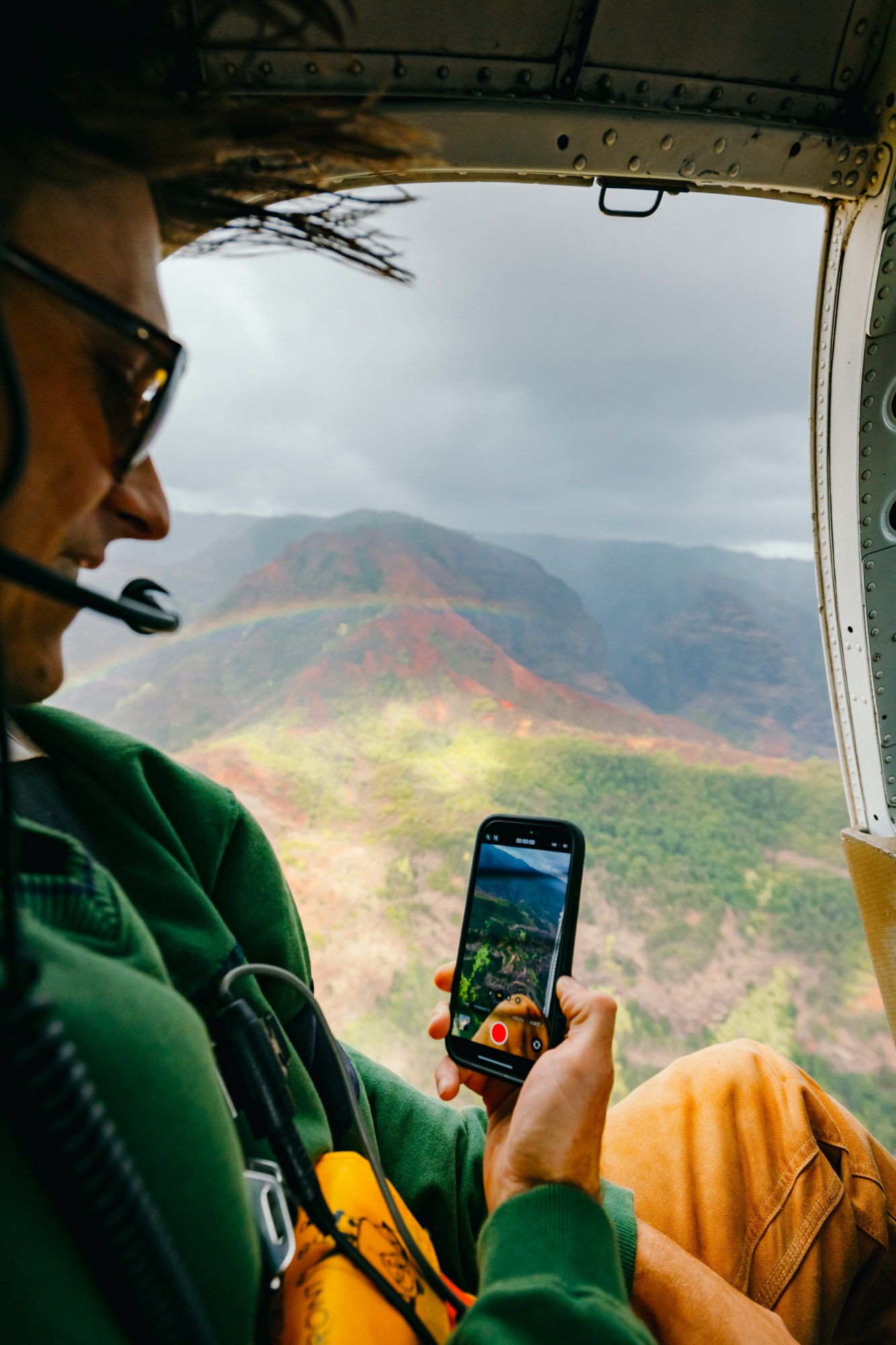 Person in helicopter using phone to capture mountain view with rainbow.